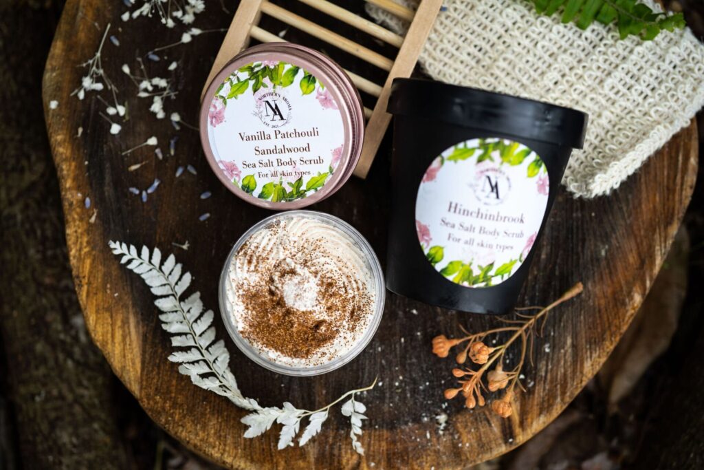 Flatlay of Northern Aroma sea salt body scrubs displayed on a rustic wooden surface. A pink-lidded Vanilla Patchouli Sandalwood scrub and a black Hinchinbrook scrub tub sit alongside an open whipped body scrub topped with cinnamon. Surrounding the products are dried botanicals, a small wooden soap rack, and a textured exfoliating cloth, creating a natural, earthy skincare aesthetic.