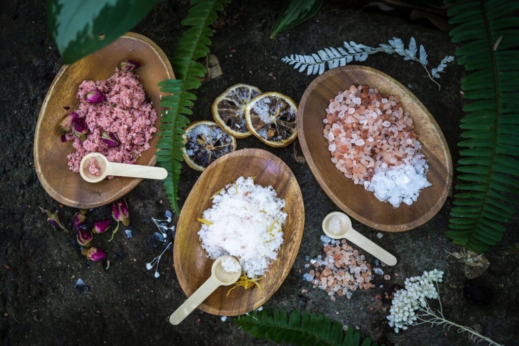Flatlay of natural bath salts displayed in wooden bowls on a dark earthy surface. One bowl holds pink rose-infused salt with dried rosebuds, another contains white salt blended with yellow petals, and a third features mixed pink Himalayan and clear salts. Small wooden scoops rest beside each bowl. Dried citrus slices, ferns, and white botanicals are arranged around the scene, creating a rustic, botanical Northern Aroma aesthetic.