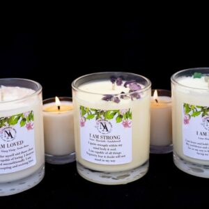 Three Northern Aroma affirmation candles—‘I Am Loved,’ ‘I Am Strong,’ and ‘I Am Brave’—displayed against a black background. Each soy candle is topped with crystals and botanicals matching its intention and scent blend. Smaller lit candles sit behind them, creating a warm, empowering, self-care atmosphere.