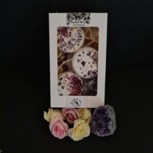 Northern Aroma ‘Shower Ritual Crystal Steamers’ gift box displayed against a dark background. Through the windowed box three white steamers topped with dried botanicals and crystals are visible. At the base of the box are pastel roses in pink, cream, and mauve, alongside a raw amethyst cluster.