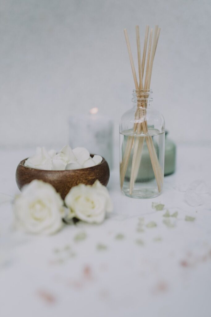 Soft, airy flatlay featuring a glass reed diffuser with natural wooden sticks, a wooden bowl filled with white rose petals, and scattered dried botanicals on a pale surface. Two white roses and a softly glowing candle in the background create a calm, elegant, spa-like atmosphere.